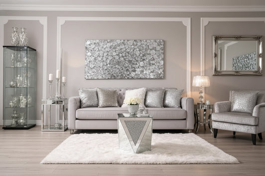 A modern living room featuring the Crystal Luxe Mirrored Accent Table, white roses in a silver vase, a light gray tufted sofa with metallic pillows, a fluffy white rug, and silver candle holders in the background.