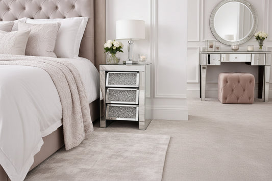 The Roma Bedside 3 Drawers adds elegant storage beside the bed, featuring a mirrored finish and topped with a glass lamp, white rose vase, and candle—perfect for softly decorated bedrooms with neutral tones.