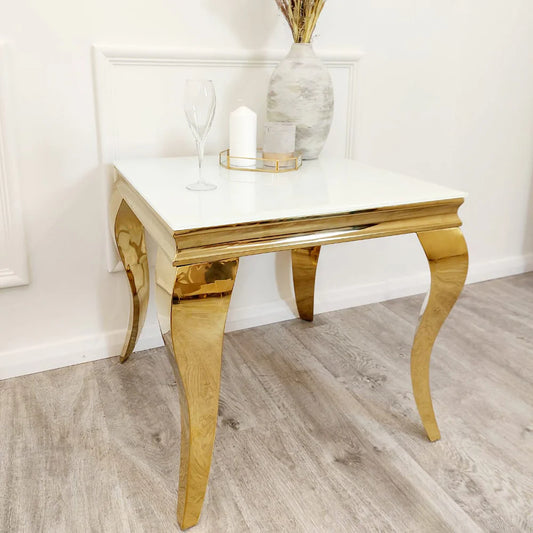 The Louis Gold Lamp Table, featuring gold curved legs and a white top, is styled with a vase of dried flowers, glass, candle, and tray—perfect against a white wall on a light wood floor.