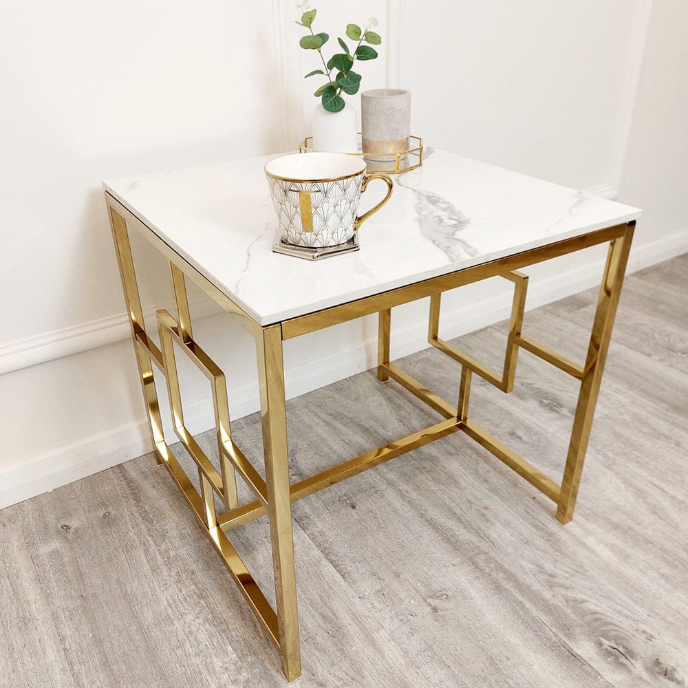 A modern side table with a white marble top and gold geometric metal legs, decorated with a patterned mug, a small candle, and a plant in a minimalist white room.