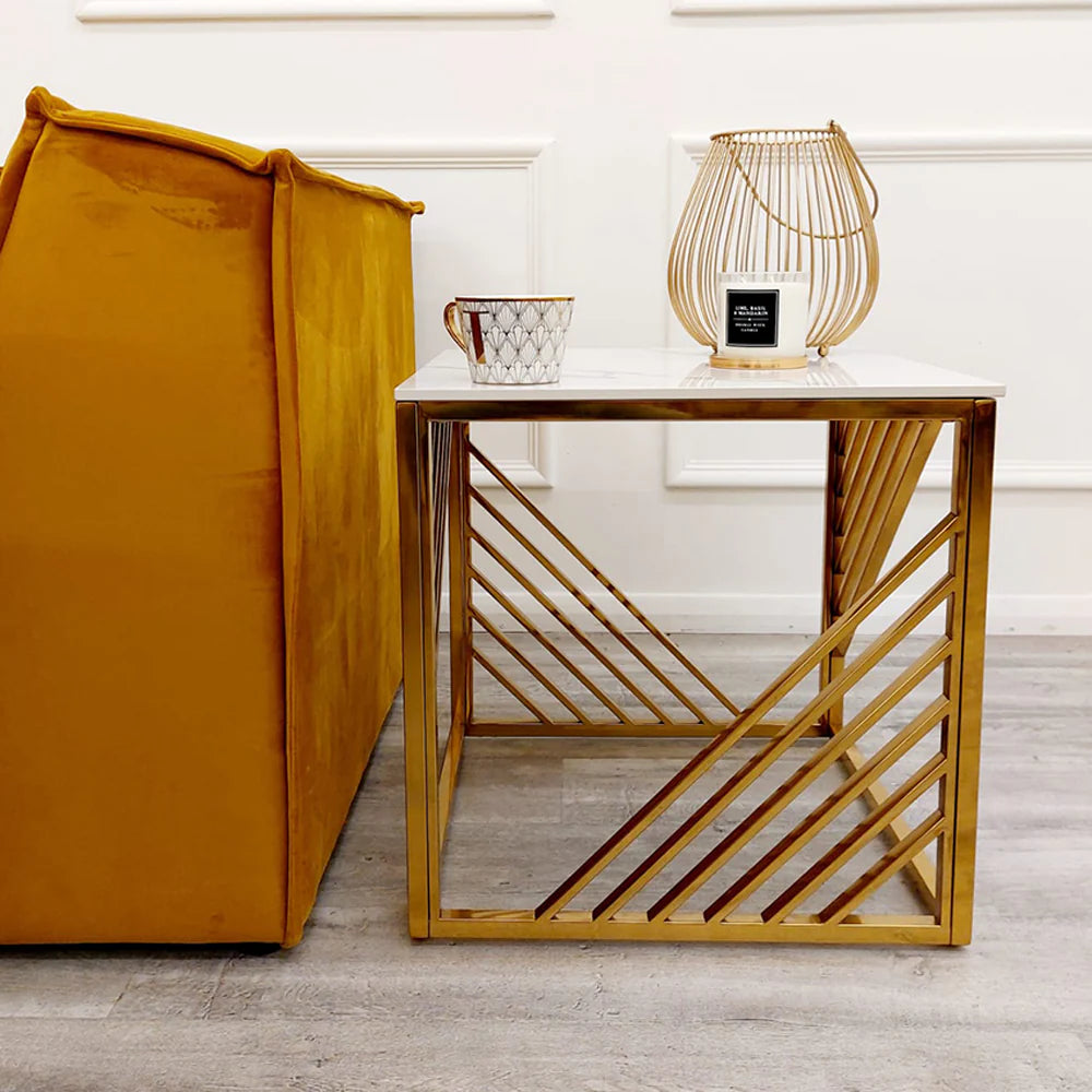 A modern Azure Gold Lamp Table with a Polar White Sintered Top stands by a mustard yellow sofa, holding a patterned mug and gold wire candle holder. The room has light wood floors and white paneled walls.