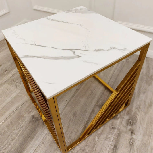 The Azure Gold Lamp Table features a polar white sintered top and a gold metal geometric base, shown on a light wood floor beside a white wall.