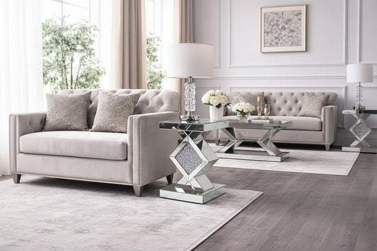 A modern living room features the Diamond Luxe Mirrored Side Table with a crystal lamp and white flowers beside a light gray tufted sofa on a patterned rug.
