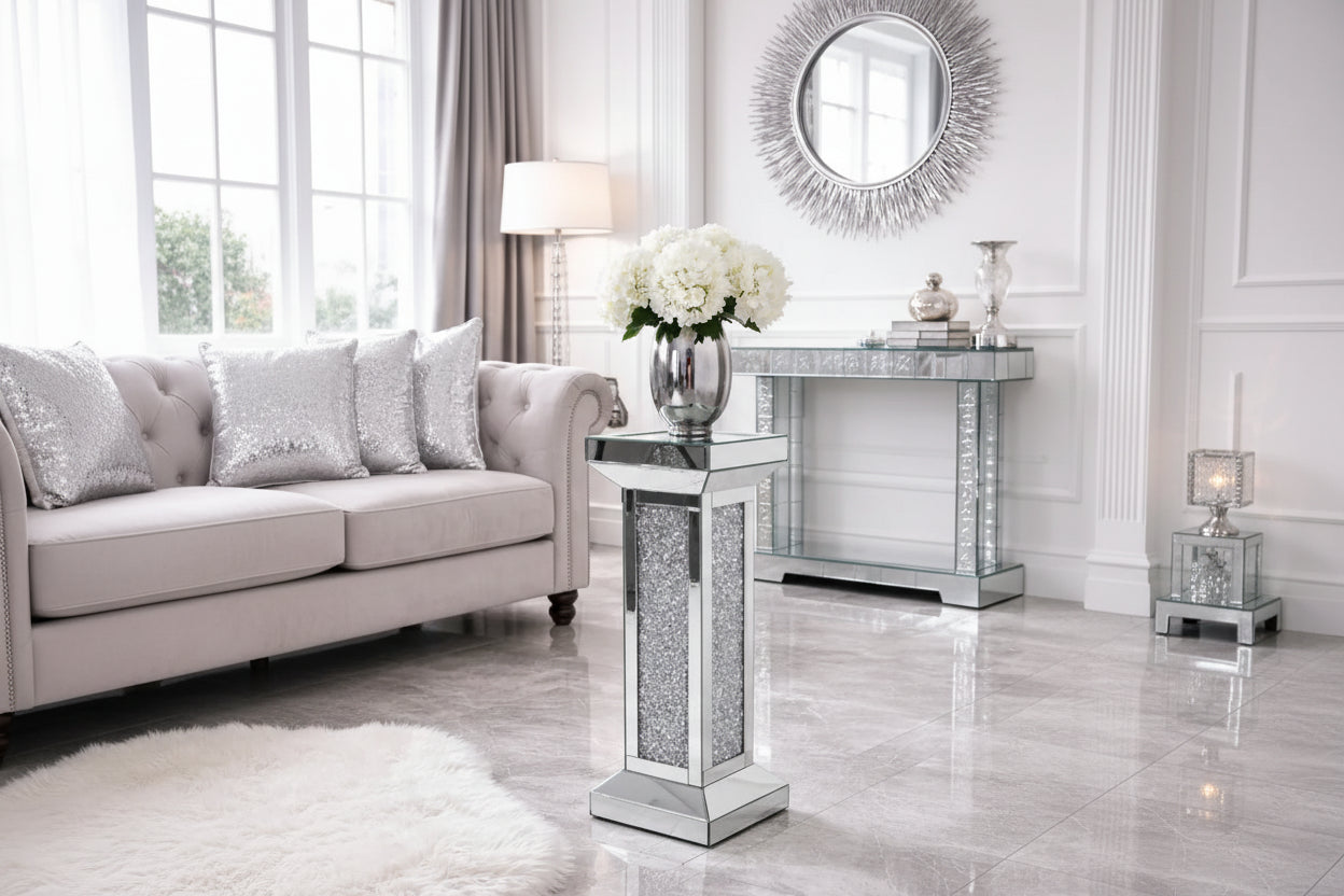 The Crystal Elegance Mirrored Pedestal Table holds a silver vase of white flowers on a marble floor in a modern living room with a tufted sofa, silver cushions, and a decorative mirror.