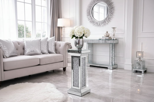 The Crystal Elegance Mirrored Pedestal Table holds a silver vase of white flowers on a marble floor in a modern living room with a tufted sofa, silver cushions, and a decorative mirror.