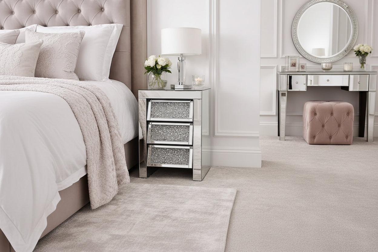 The Roma Bedside 3 Drawers adds elegant storage beside the bed, featuring a mirrored finish and topped with a glass lamp, white rose vase, and candle—perfect for softly decorated bedrooms with neutral tones.
