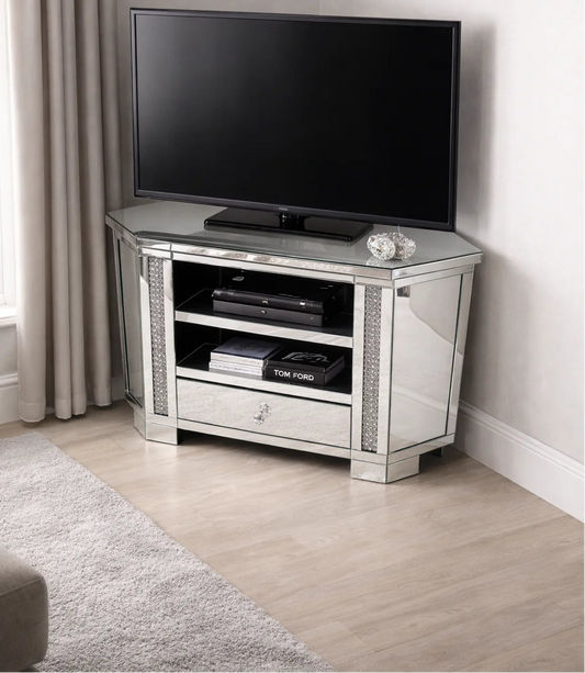 A flat-screen TV rests on the Roma Corner Mirrored TV Unit with a glass drawer and shelves for electronics, beside cream curtains and a beige carpet on light wood flooring—a stylish, space-saving TV solution.