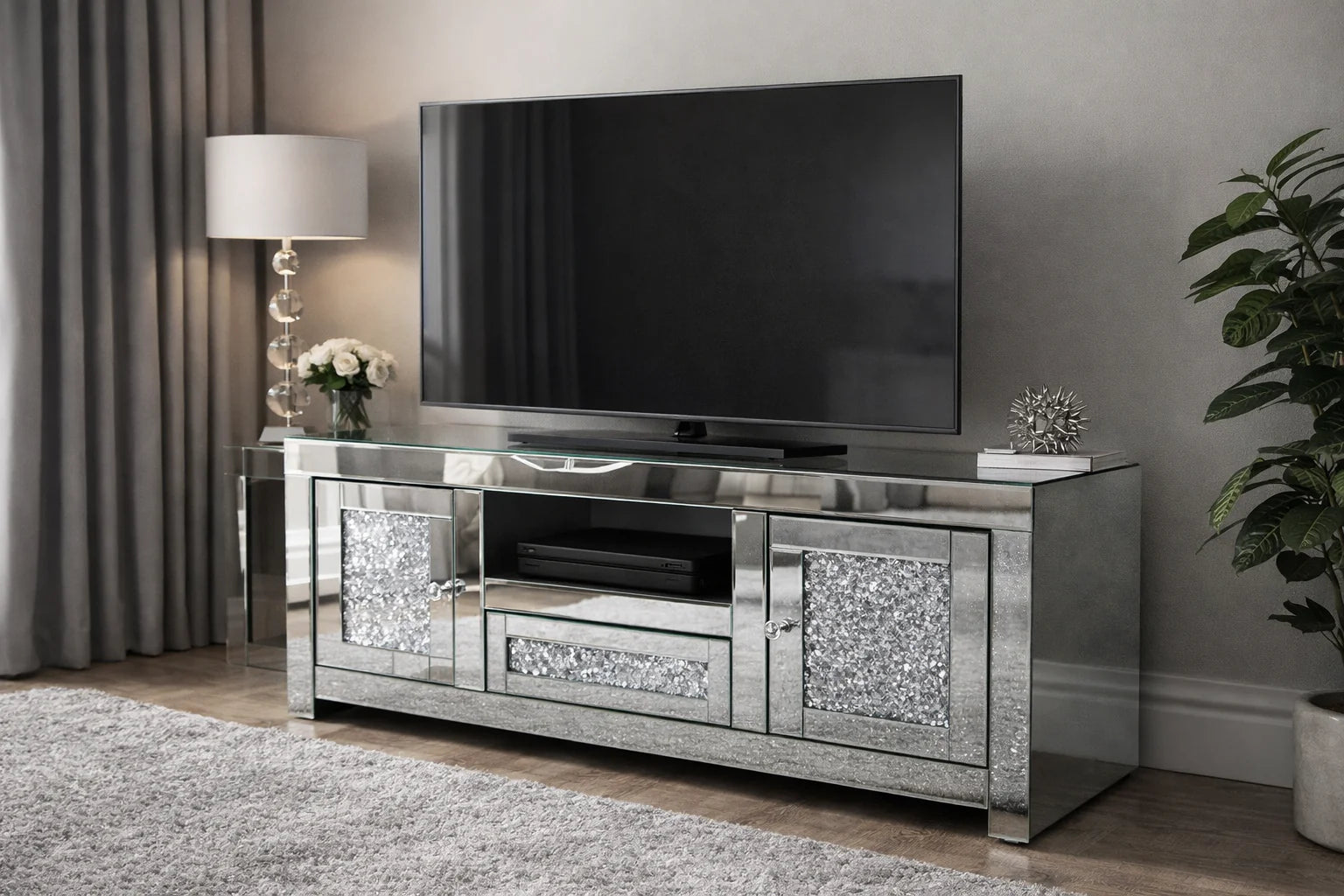 A modern living room with a large flat-screen TV on the Roma Mirrored TV Unit | Crushed Diamond TV Stand, adorned with a lamp, white flowers, a plant, and silver accent. Curtains and a rug complete this cozy, stylish space.