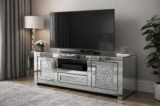 A modern living room with a large flat-screen TV on the Roma Mirrored TV Unit | Crushed Diamond TV Stand, adorned with a lamp, white flowers, a plant, and silver accent. Curtains and a rug complete this cozy, stylish space.