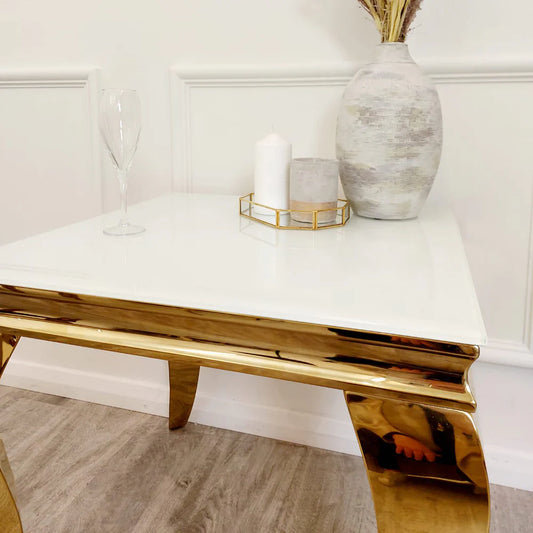The Louis Gold Lamp Table features a glossy white top and shiny gold legs, styled with a wine glass, gold tray with white candles, and a textured white vase with dried stems, all set before a white paneled wall.