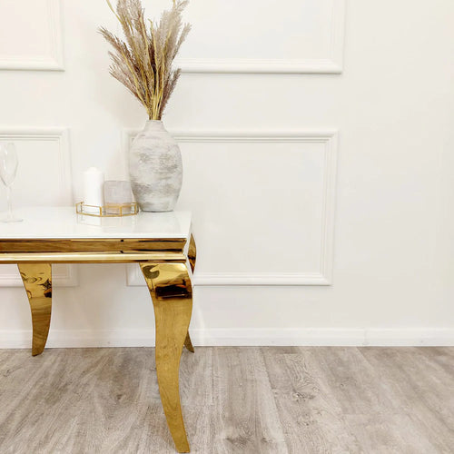 A modern room showcases the Louis Gold Lamp Table with gold legs and a white top, styled with a white vase of dried grasses, a candle, and a glass. The light wood floor and white paneled wall add to the elegant decor.