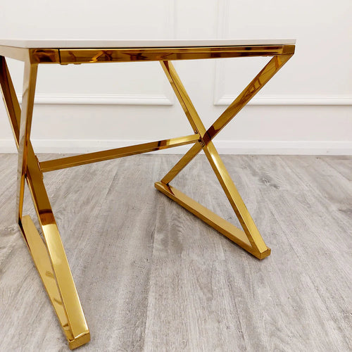 A close-up of the Zion Gold Lamp Table with a polar white sintered top and shiny gold X-shaped legs, standing on a light gray wood floor against a white wall with trim.