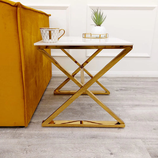 The Zion Gold Lamp Table with Polar White Sintered Top features a gold metal frame and sits by a mustard yellow sofa, holding a geometric mug, glass container, and potted plant on the light wood floor.